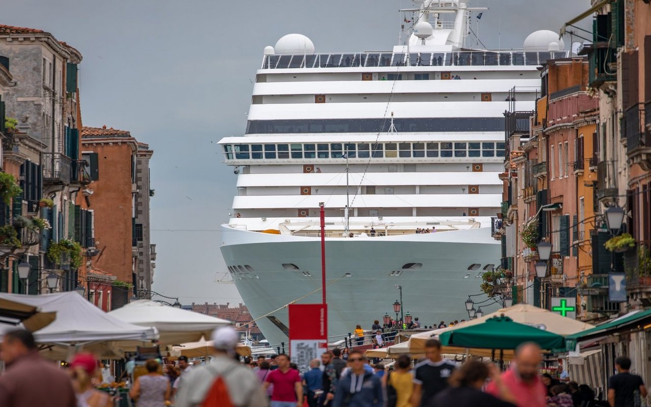 Fast bedrohlich ragt das Kreuzfahrtschiff über die Hafenstadt auf. Ein solches Schiff kann tausende Passagiere gleichzeitig an Land bringen, dabei Infrastruktur und öffentliche Räume überlasten und zudem spürbare Umweltbelastungen in der Region verursachen.
